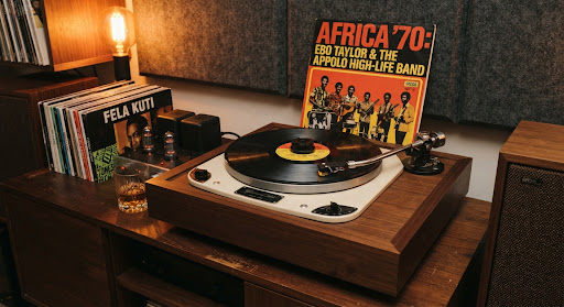 A vintage Garrard 301 turntable with high-end tonearm spinning an original 1970s Ghanaian highlife vinyl record on dark walnut console. Warm ambient filament bulb glow casts amber reflections across sleeve art featuring bold West African typography, surrounded by acoustic wool lounge panels.